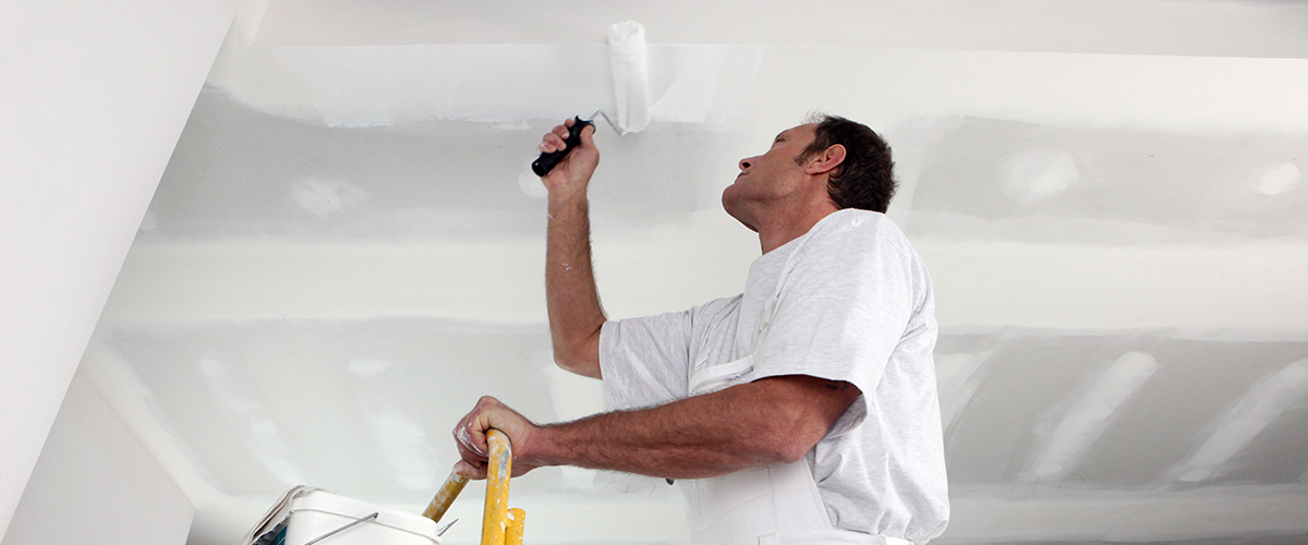 A painter on a ladder, using a roller to paint a white ceiling, with a paint tray and tools beside him.