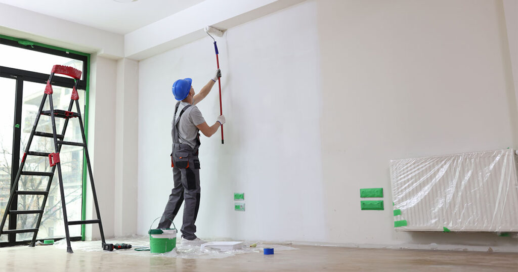 A painter wearing a blue helmet and gray work clothes is using a roller brush on a long handle to paint a large white wall in a room.