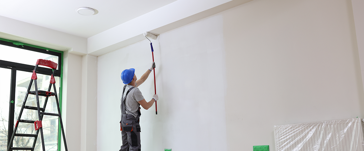 A painter wearing a blue helmet and gray work clothes is using a roller brush on a long handle to paint a large white wall in a room.