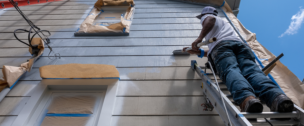 A worker on a ladder painting the exterior of a house, with windows and other areas masked off with paper and tape.