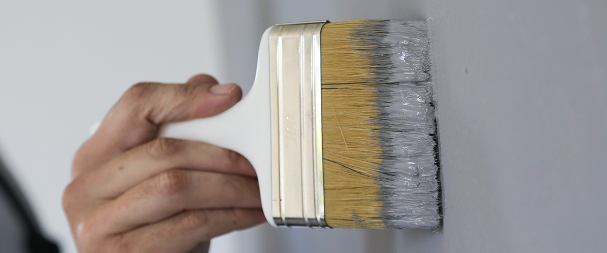 Close-up of a hand holding a paintbrush applying gray paint to a wall, showcasing interior wall painting process.