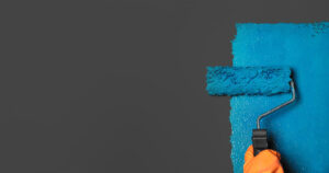 A hand in an orange glove uses a paint roller to apply blue paint on a gray wall.