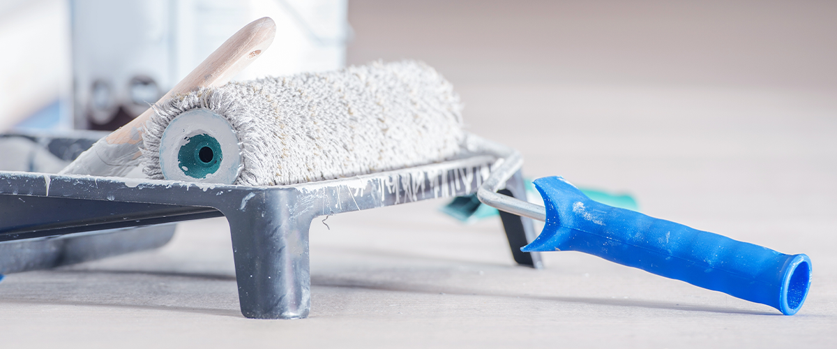 Close-up of aa paint roller with blue handle resting on a tray, with a white paint can in the background, prepared for interior painting.