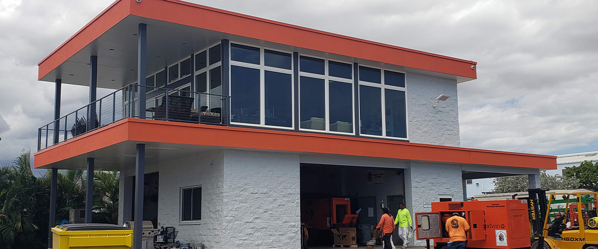 Commercial Painting In Wahiawa, HI. Modern industrial building with white brick exterior and orange trim, featuring large windows and an upper balcony. Workers and equipment visible.