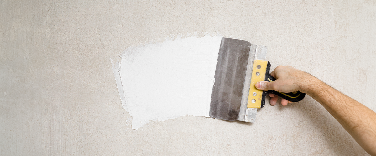 Hand using a drywall taping knife to apply white joint compound on a beige wall for a smooth finish, home improvement, stucco repair.