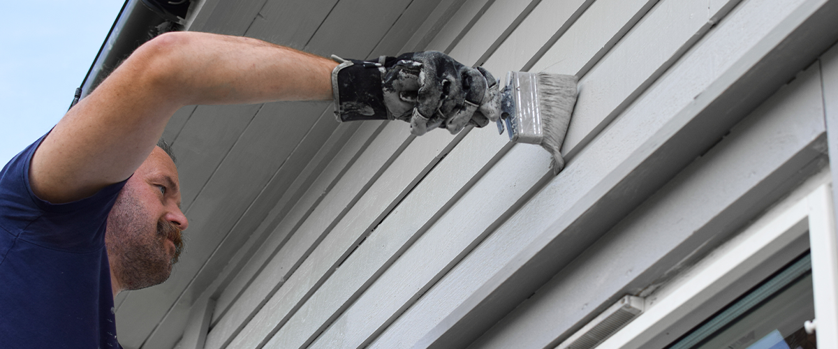 Man doing an exterior painting job in Wahiawa, with a brush, applying fresh white paint to the wooden siding.