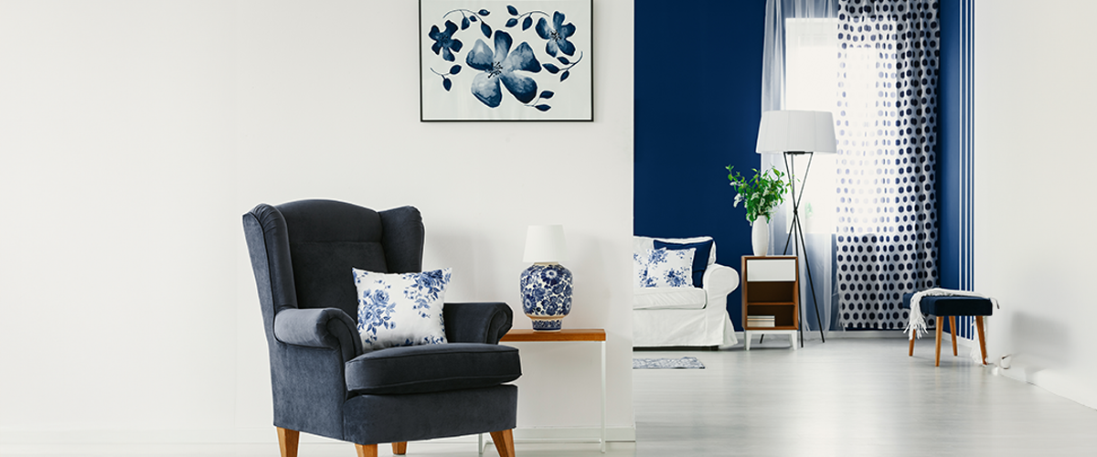 Modern living room with a white and navy blue color scheme, featuring a dark blue armchair, floral cushions, a matching lamp, and a cozy white sofa against a navy accent wall.