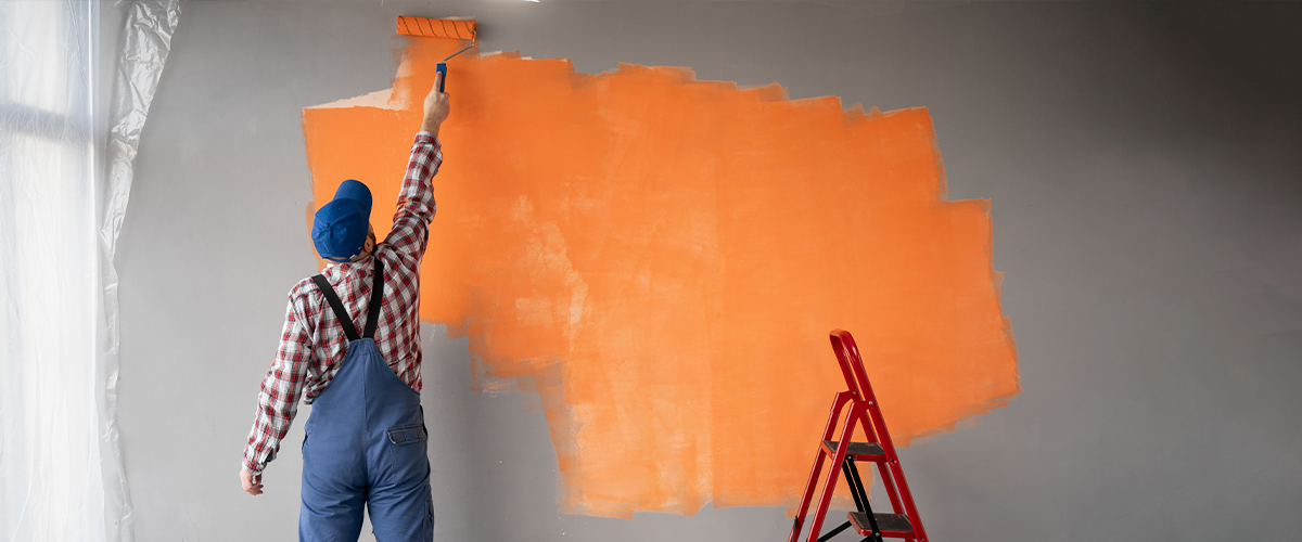 Painter in blue overalls and a cap applying bright orange paint on a grey wall with a roller, while a red ladder stands nearby.