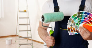 Painter in overalls holding a paint roller and a color palette in a prepared room with ladder and painting supplies.