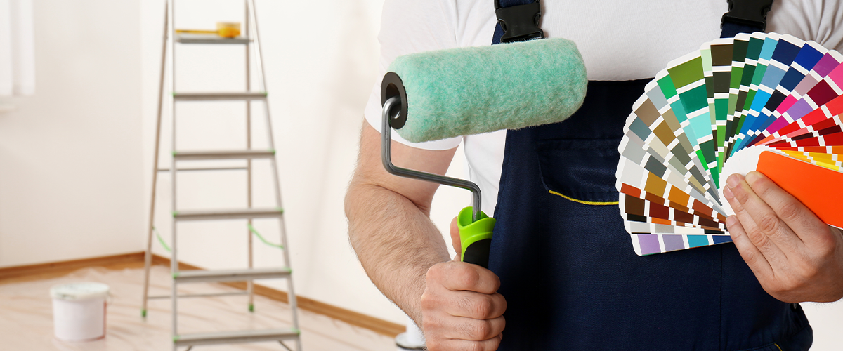 Painter in overalls holding a paint roller and a color palette in a prepared room with ladder and painting supplies.