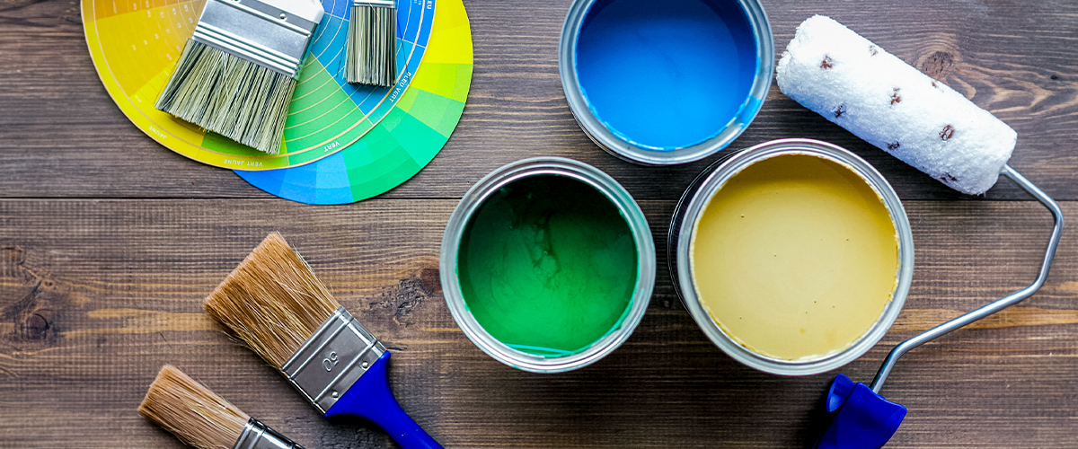 Top view of a set of painting tools on a wooden surface, including open cans of blue, green, and yellow paint, several paintbrushes, a color wheel, and a paint roller.