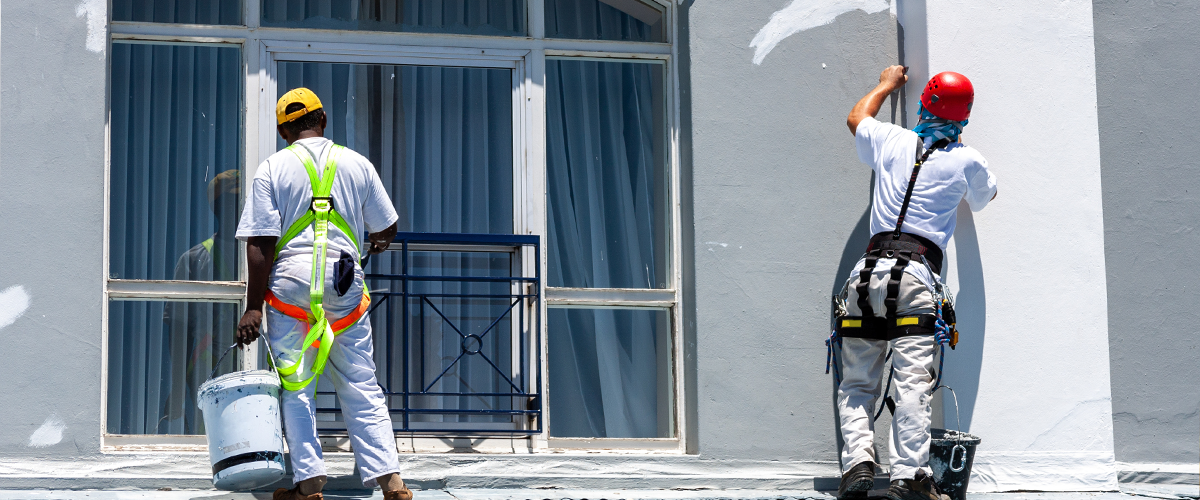 Two workers doing a commercial painting project in Wahiawa, while wearing safety harnesses and helmets. One worker holds a paint bucket, and both are on a corrugated roof.