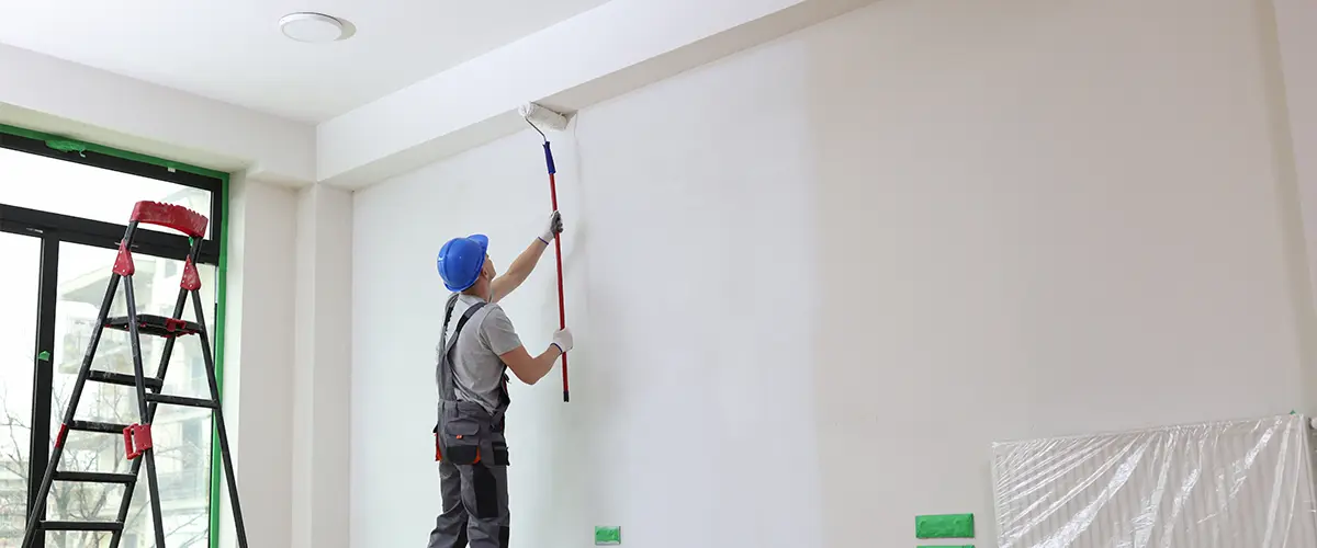 Construction worker painting interior wall with roller, wearing hard hat and overalls, ladder and tools visible in modern renovation.