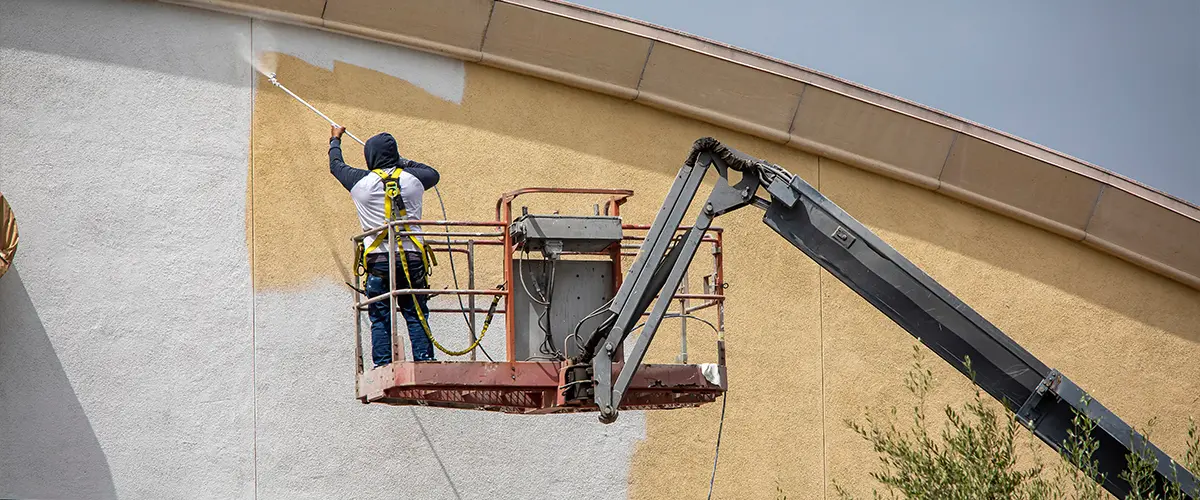 Worker painting exterior wall using spray gun on construction lift, commercial building painting project.