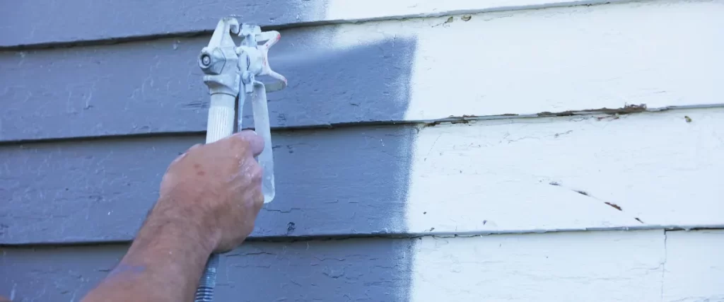 A professional painter using a spray gun to apply a fresh coat of gray paint on an old wooden house exterior, covering the previous white color.