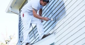 A professional painter standing on a ladder, applying fresh gray paint to the worn wooden siding of a house using a paintbrush