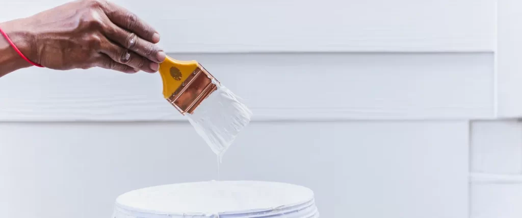 A person dipping a paintbrush into a bucket of white paint, preparing to apply a fresh coat on the textured siding of a house