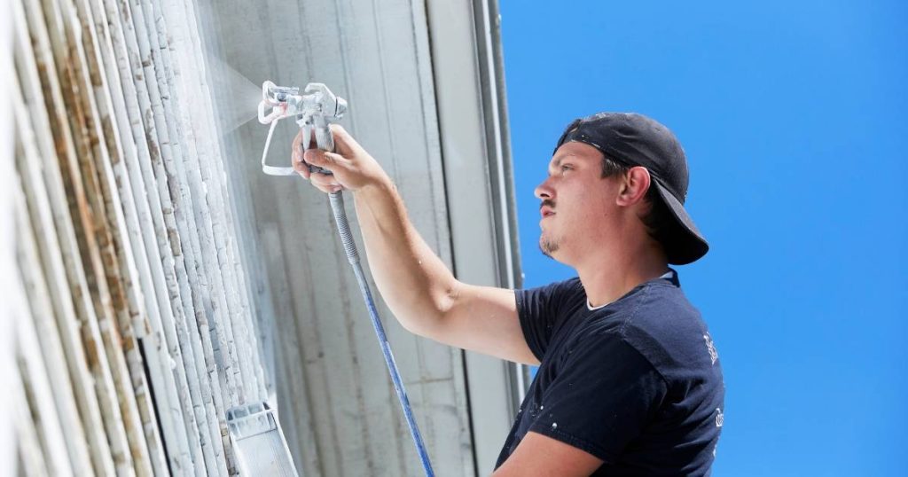 The Best Time of Year to Paint a Home in Mililani: Weather Considerations 3 Man using a paint sprayer on a tall building under a clear blue sky