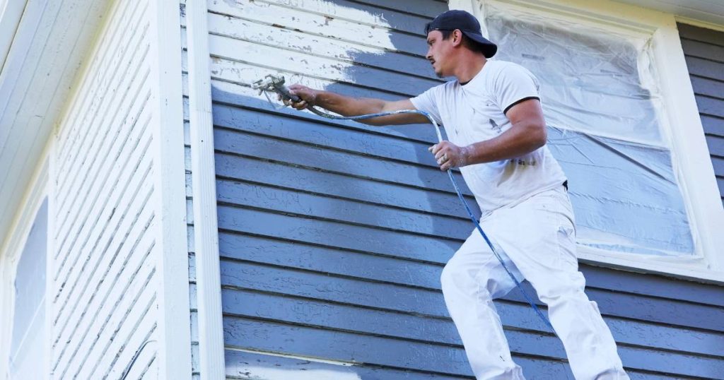 Why Hawaii Homes Need Regular Exterior Repainting: Sun, Salt & Rain Damage Explained 4 Exterior painter applying fresh white paint with a sprayer on blue siding