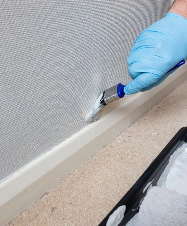 Baseboard Painting 1 A person wearing blue protective gloves uses a small brush for baseboard painting along a textured wall with masking tape for clean edges.
