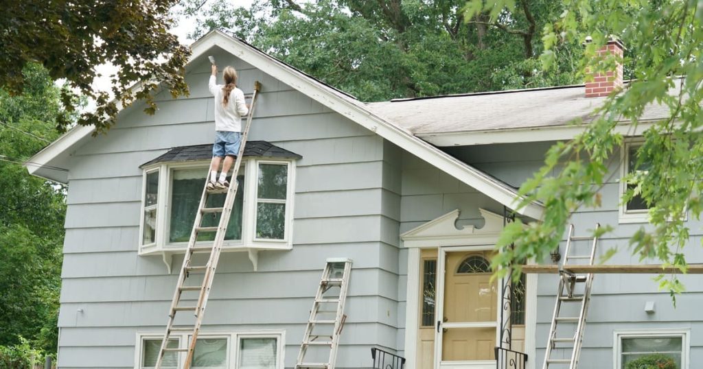 A person standing on a tall wooden ladder while repainting house exterior trim and siding on a light blue two-story home.