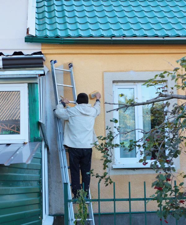 Top 8 Exterior Painting Companies In Pearl City, HI 1 A professional painter on a ladder applying a fresh coat of tan paint to the side of a house, showcasing the quality work of exterior painting companies in Pearl City, HI.
